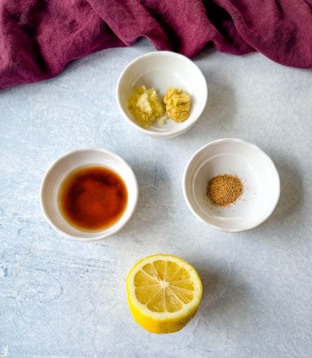Flat lay of individual remoulade sauce ingredients in small white bowls, including Dijon mustard, minced garlic, hot sauce, Creole seasoning, and a halved lemon, set against a light blue background.