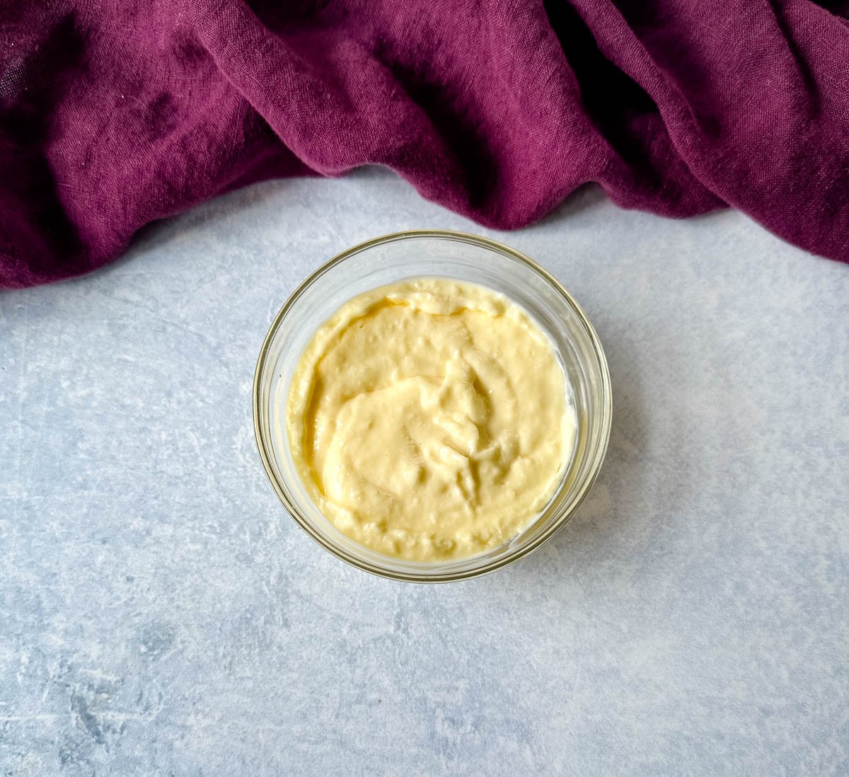 Overhead shot of creamy Louisiana-style remoulade sauce in a small glass bowl on a light surface, with a deep purple kitchen towel in the background.