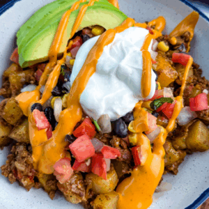 Bowl of loaded taco potatoes topped with avocado slices, black beans, corn, tomatoes, sour cream, and creamy orange sauce drizzle.