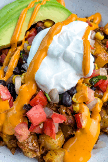 Bowl of loaded taco potatoes topped with avocado slices, black beans, corn, tomatoes, sour cream, and creamy orange sauce drizzle.