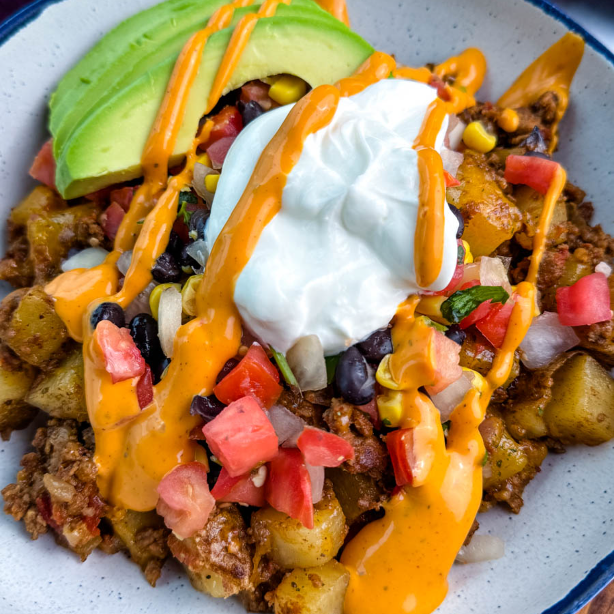 Bowl of loaded taco potatoes topped with avocado slices, black beans, corn, tomatoes, sour cream, and creamy orange sauce drizzle.