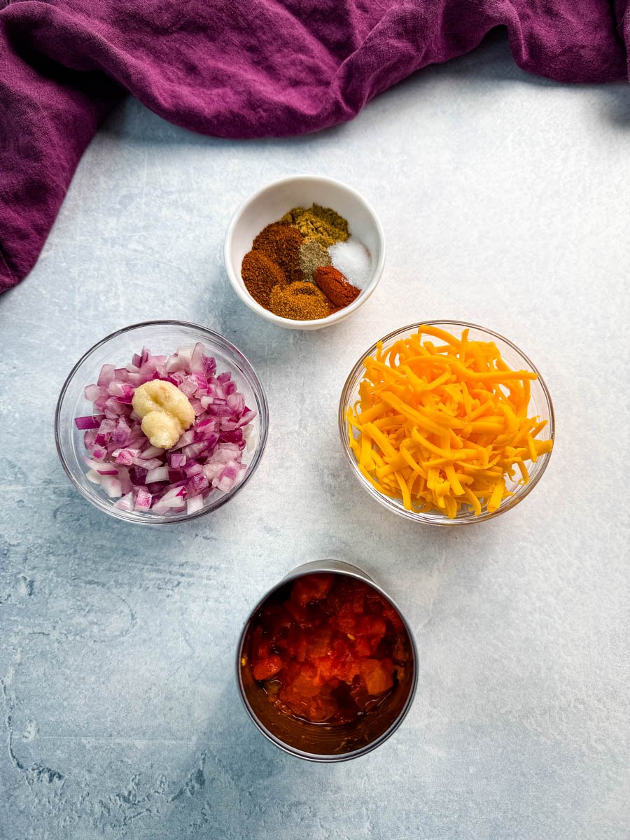 Flat lay of prepared ingredients: diced red onion, minced garlic, shredded cheddar cheese, canned diced tomatoes with chilies, and a bowl of taco seasoning.