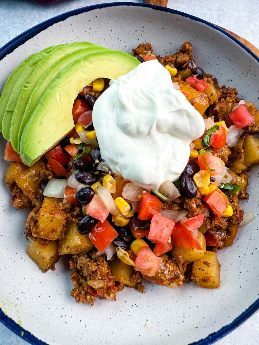 Bowl filled with taco-seasoned potatoes and ground meat, topped with sour cream, avocado, corn, black beans, and fresh diced veggies for a colorful, one-pot dinner.