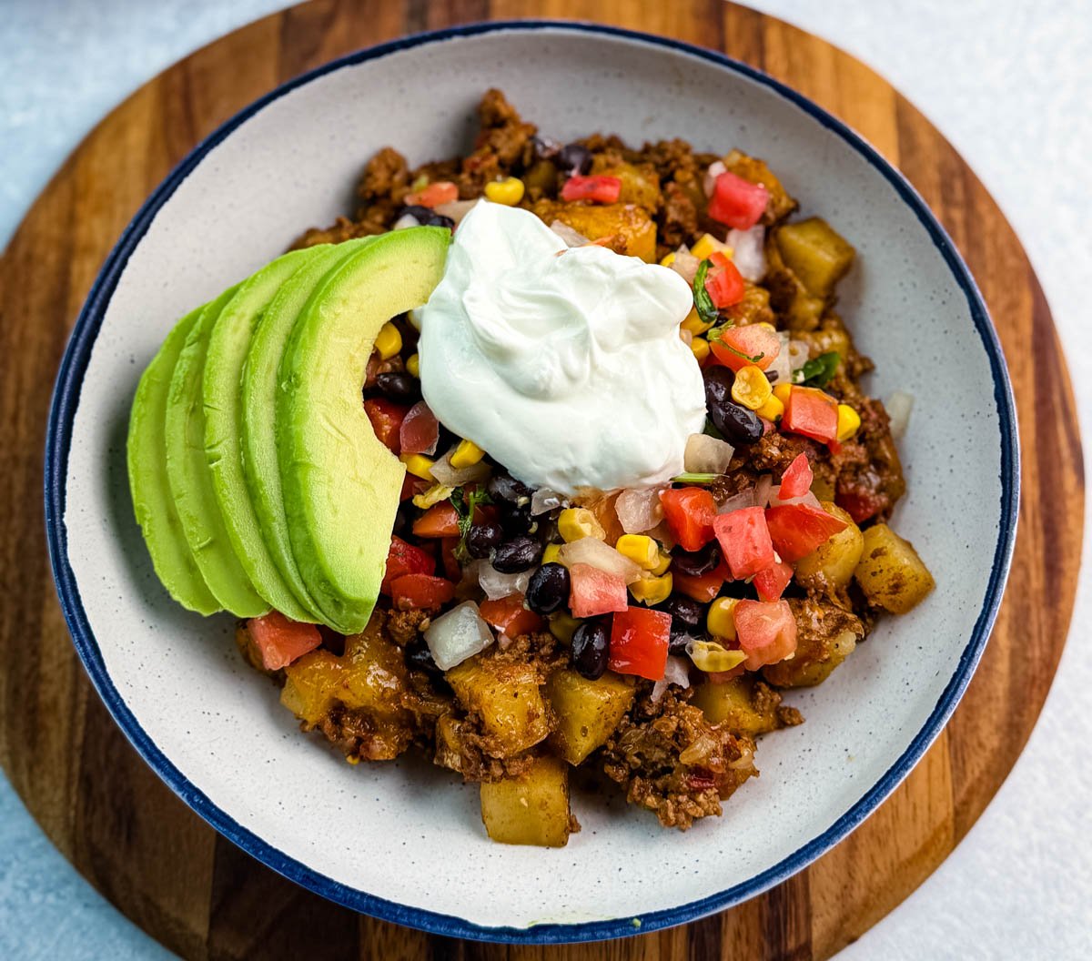 Center view of a hearty taco potato bowl loaded with seasoned meat, potatoes, avocado, and toppings, styled on a round white plate with a navy rim.