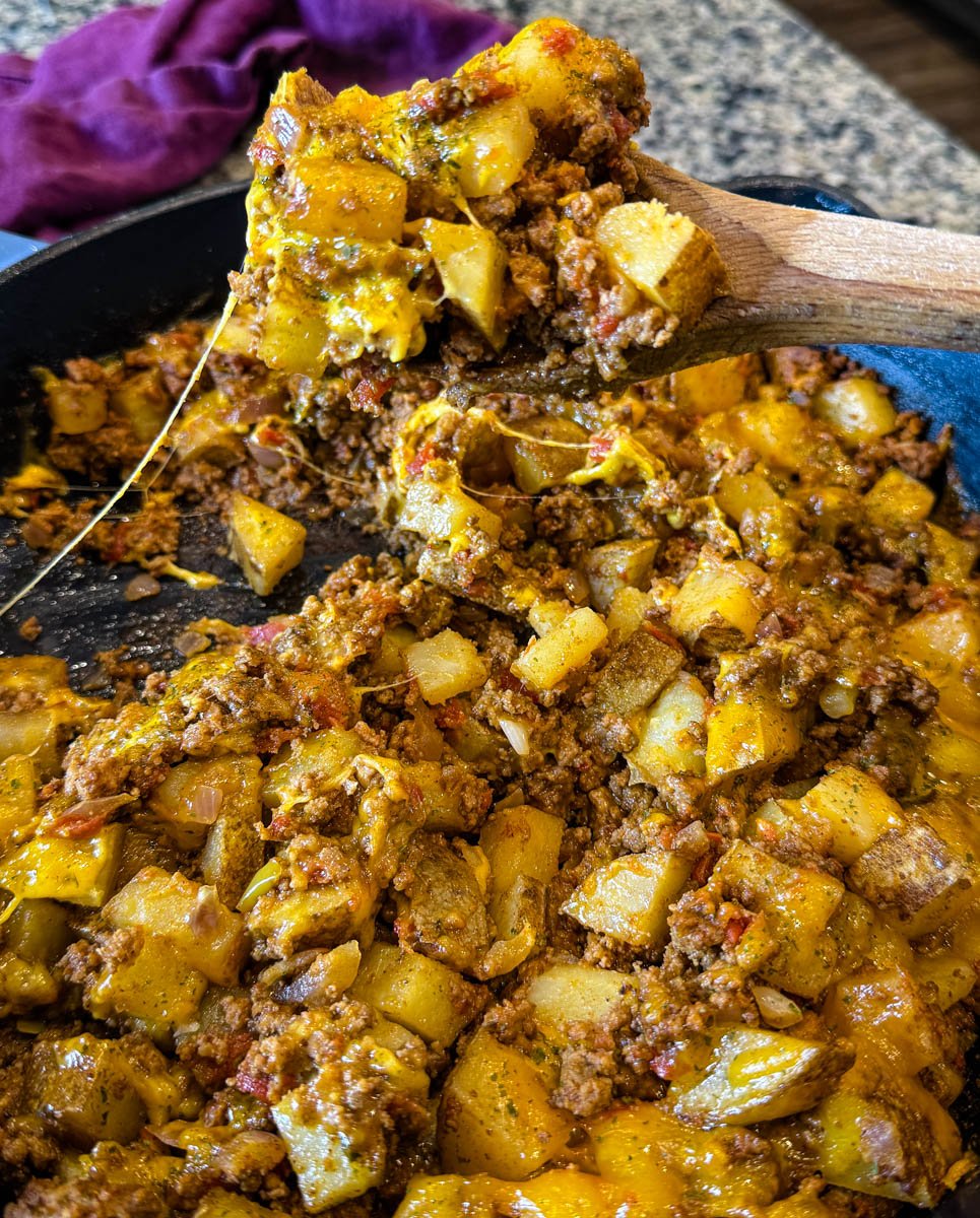 Close-up of a wooden spoon scooping cheesy taco potato mixture straight from a hot skillet—melted cheese and savory filling in every bite.