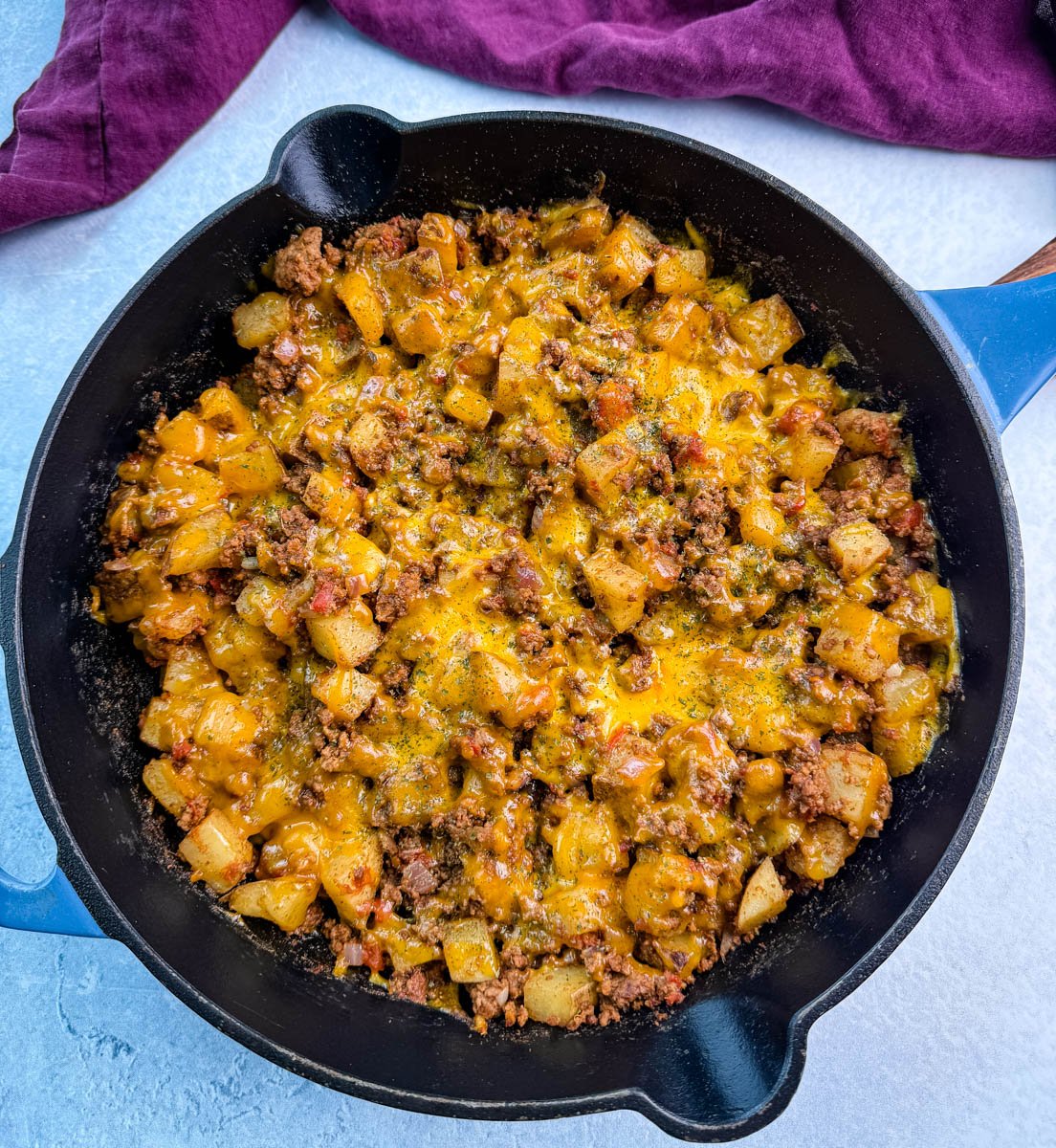 Full view of a cast iron skillet packed with cheesy taco potatoes, topped with herbs, sitting on a light blue surface with a purple kitchen towel nearby.
