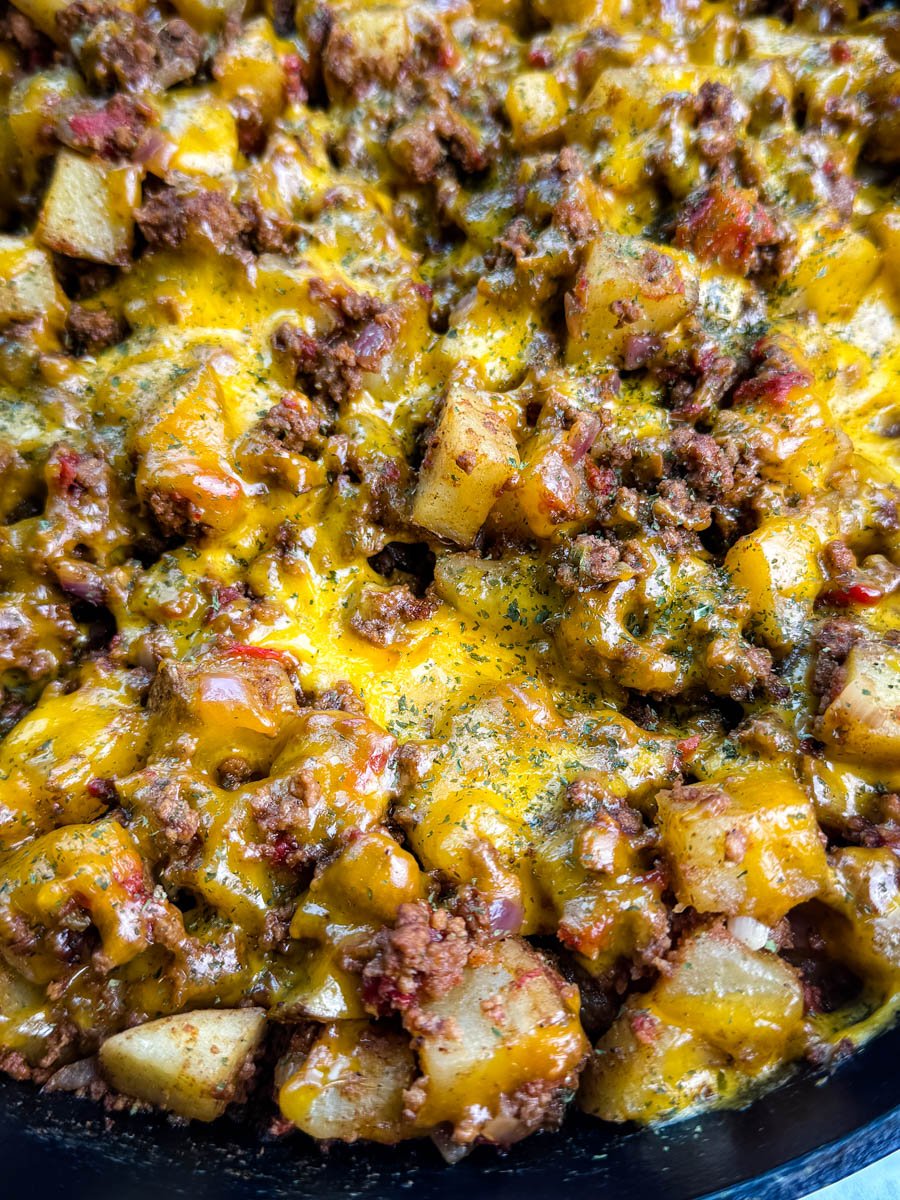 Extreme close-up of cheesy taco potatoes with ground beef, diced tomatoes, and melted cheddar, garnished with herbs in a cast iron skillet.