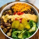 Top-down view of a Big Mac bowl with roasted potatoes, ground beef, shredded cheddar, chopped tomatoes, lettuce, pickles, and a drizzle of homemade sauce, served on a wooden board.