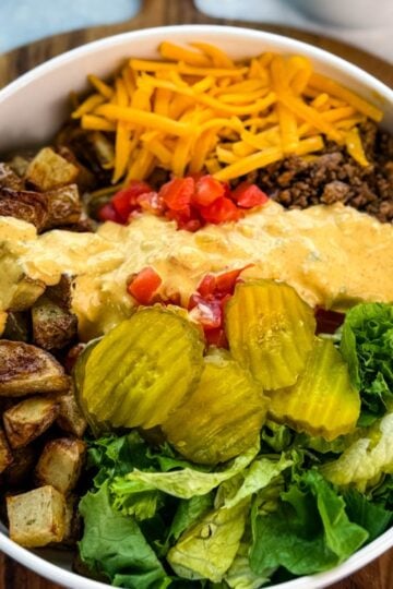 Top-down view of a Big Mac bowl with roasted potatoes, ground beef, shredded cheddar, chopped tomatoes, lettuce, pickles, and a drizzle of homemade sauce, served on a wooden board.
