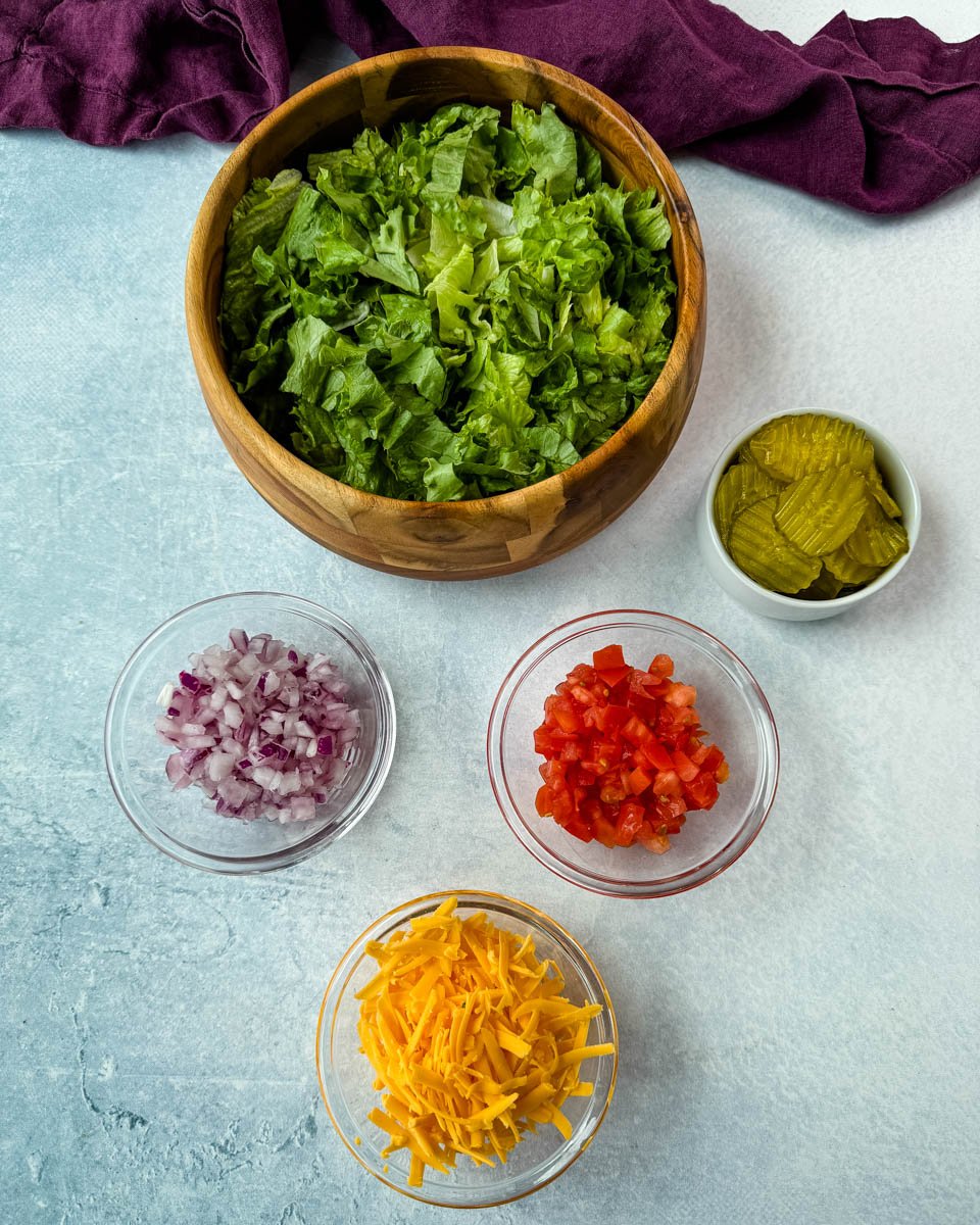 Ingredient prep for Big Mac bowls, featuring shredded lettuce in a wooden bowl, and smaller glass bowls of diced red onion, tomatoes, shredded cheddar cheese, and sliced pickles.