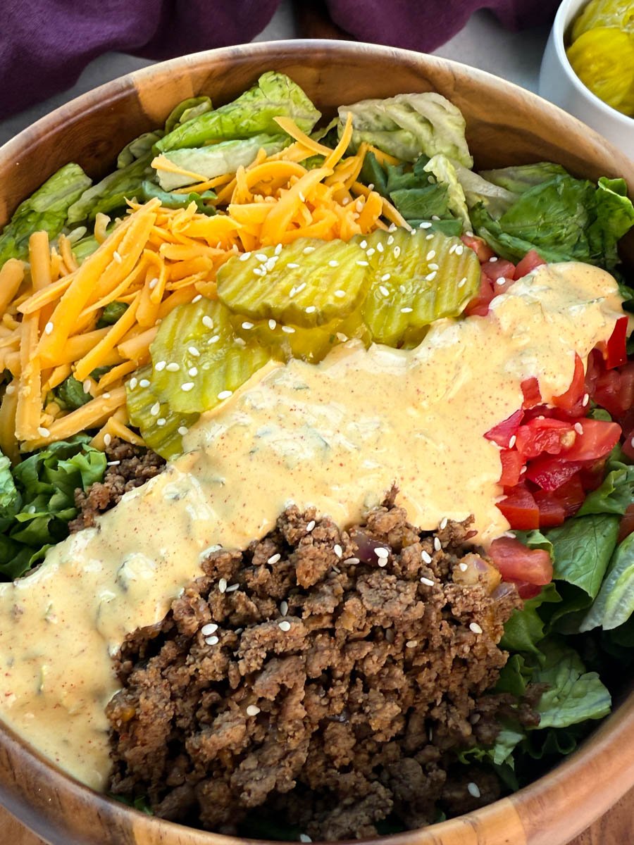 Close-up of Big Mac salad featuring a generous layer of pickles, shredded cheddar cheese, fresh lettuce, and ground beef with a creamy sauce drizzle and sesame seed garnish.