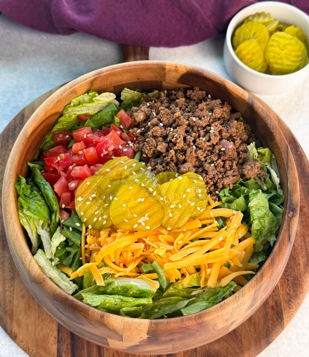 Overhead view of a Big Mac salad loaded with seasoned beef, tomatoes, shredded lettuce, cheddar cheese, and pickles, served in a wooden bowl with sesame seeds on top.