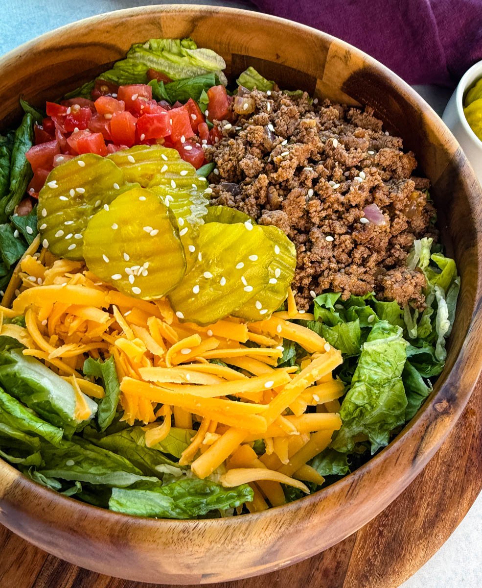 Detailed close-up shot showing textures of shredded cheddar, lettuce, pickles, and ground beef in a Big Mac salad topped with sauce and sesame seeds.