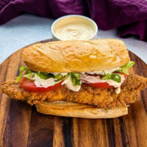 Close-up of a finished catfish po’ boy sitting on a wooden cutting board. The sandwich is stuffed with crispy golden fish, lettuce, tomato, and drizzled with creamy sauce. A bowl of extra sauce is blurred in the background.