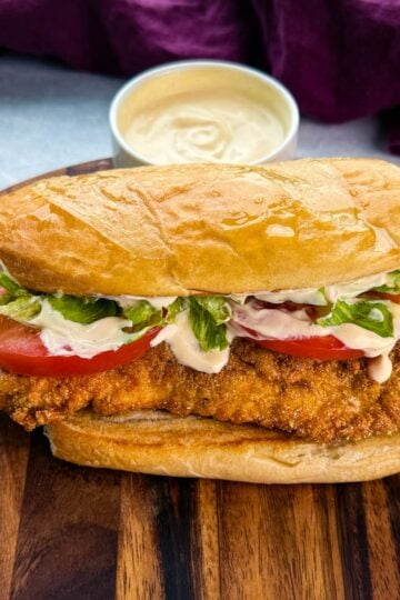 Close-up of a finished catfish po’ boy sitting on a wooden cutting board. The sandwich is stuffed with crispy golden fish, lettuce, tomato, and drizzled with creamy sauce. A bowl of extra sauce is blurred in the background.