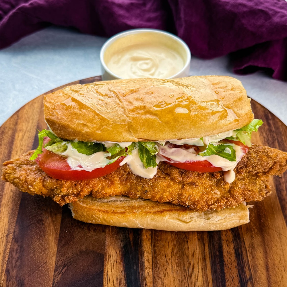 Close-up of a finished catfish po’ boy sitting on a wooden cutting board. The sandwich is stuffed with crispy golden fish, lettuce, tomato, and drizzled with creamy sauce. A bowl of extra sauce is blurred in the background.