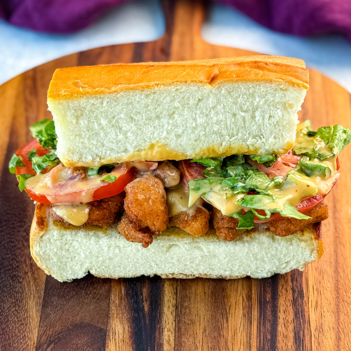 Centered image of a crispy chicken po' boy sandwich on a cutting board, showcasing layers of fried chicken, tomato, lettuce, and sauce, surrounded by chips.