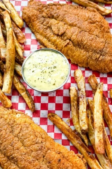 Golden fried catfish served with hand-cut fries and a side of creamy tartar sauce, all arranged on red and white checkered paper in a black tray.