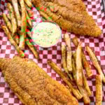 Golden fried catfish served with hand-cut fries and a side of creamy tartar sauce, all arranged on red and white checkered paper in a black tray.