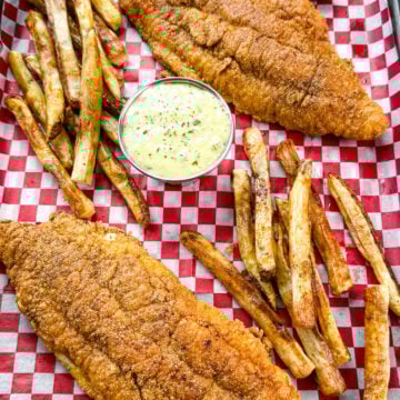 Golden fried catfish served with hand-cut fries and a side of creamy tartar sauce, all arranged on red and white checkered paper in a black tray.