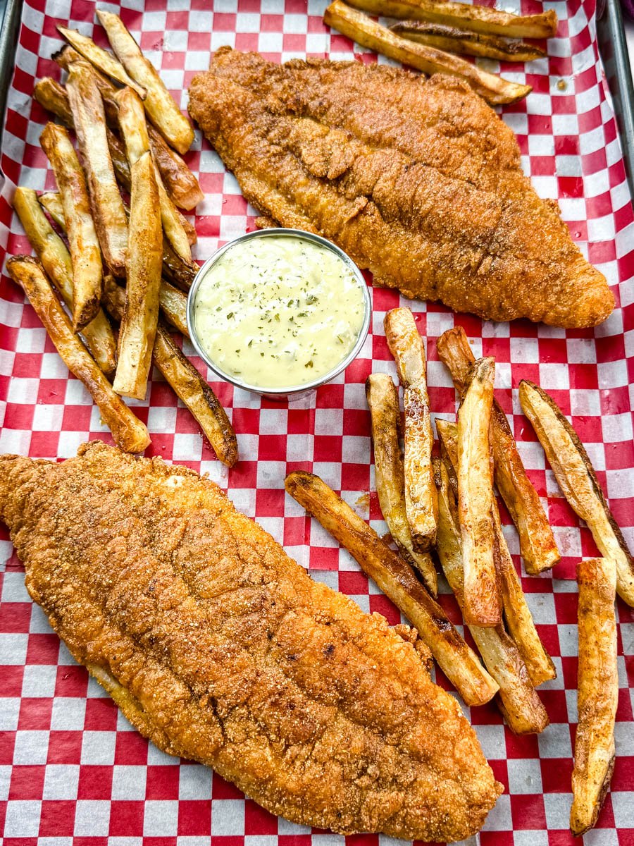 Golden fried catfish served with hand-cut fries and a side of creamy tartar sauce, all arranged on red and white checkered paper in a black tray.