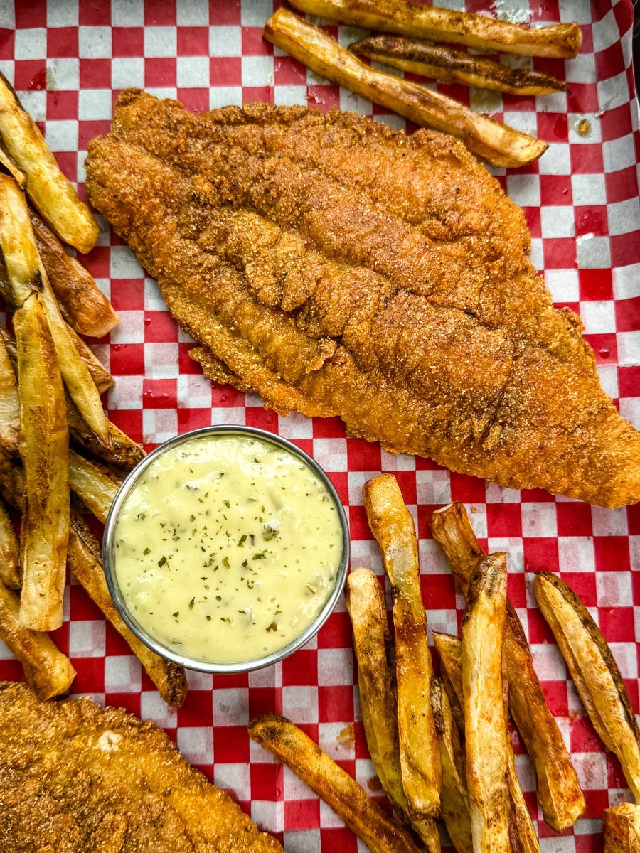 Overhead view of a crispy catfish fillet surrounded by fries and a cup of tartar sauce, all laid out on red and white checkered paper.