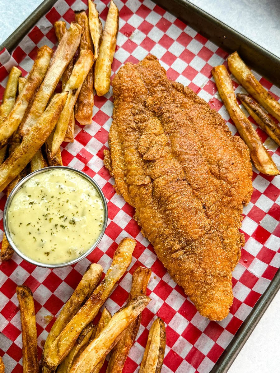Fried catfish and crispy fries on a serving tray with a bowl of tartar sauce, styled on checkered deli paper with a bright, Southern fish fry feel.