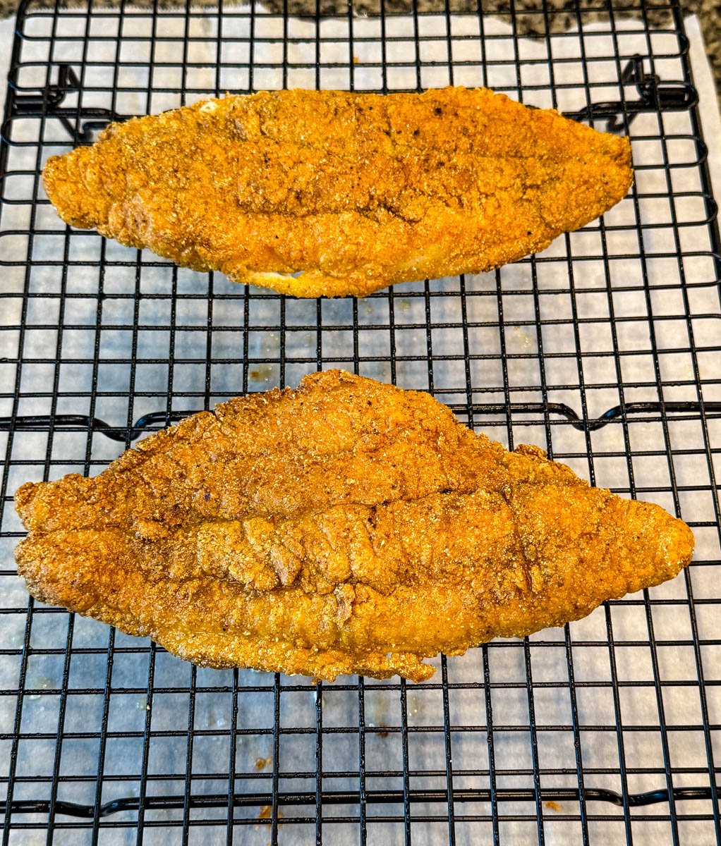 Two golden, crispy fried catfish fillets resting on a wire rack for cooling or draining, placed over parchment paper.