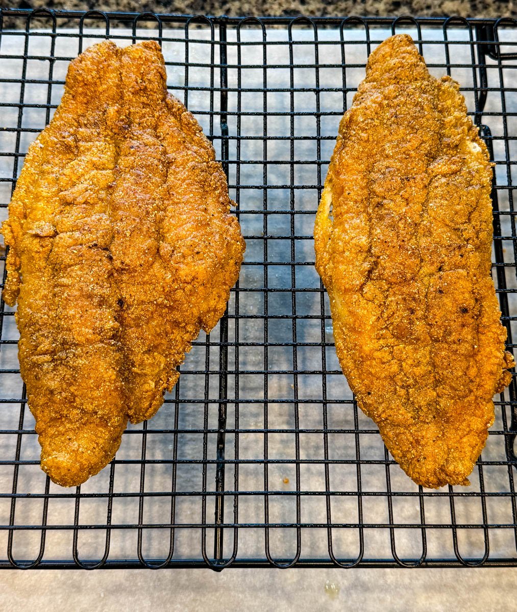 Two golden, crispy fried catfish fillets resting on a wire rack for cooling or draining, placed over parchment paper.