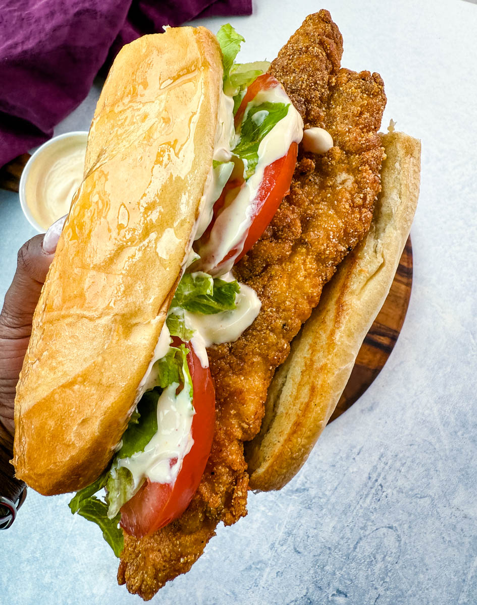 Overhead view of po' boy sandwich ingredients arranged on a round wooden cutting board. The setup includes shredded lettuce, three slices of ripe tomato, a small bowl of creamy sauce, and a large loaf of French bread. A purple cloth napkin is placed above the board on a light background.