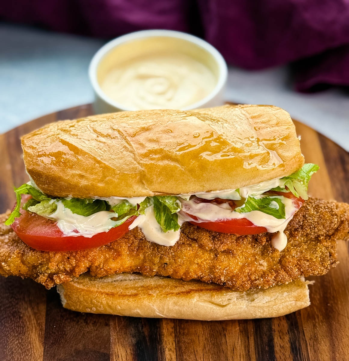Close-up of a finished catfish po’ boy sitting on a wooden cutting board. The sandwich is stuffed with crispy golden fish, lettuce, tomato, and drizzled with creamy sauce. A bowl of extra sauce is blurred in the background.
