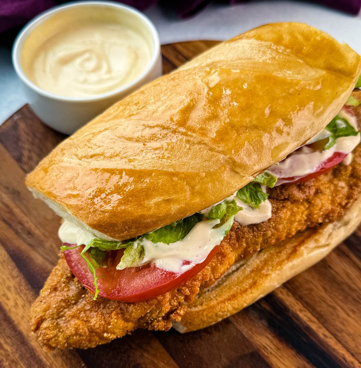 A different angle of the catfish po’ boy showing the glistening toasted top bun and sandwich fillings peeking out: shredded lettuce, tomatoes, and sauce over a crisp fillet.