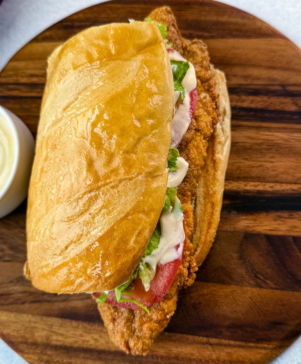 Overhead shot of the sandwich with a shiny top bun and toppings visible. A bowl of dipping sauce is placed beside it on a wooden board.
