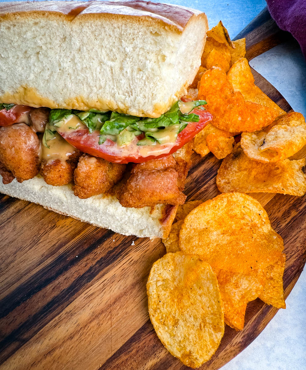 A close-up of a crispy chicken po' boy sandwich on a wooden board, topped with lettuce, tomato, and creamy sauce, served with seasoned potato chips on the side.