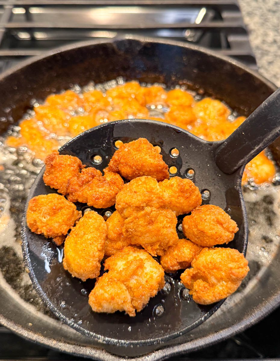 A slotted spoon lifting crispy fried chicken bites out of hot oil in a cast iron skillet.
