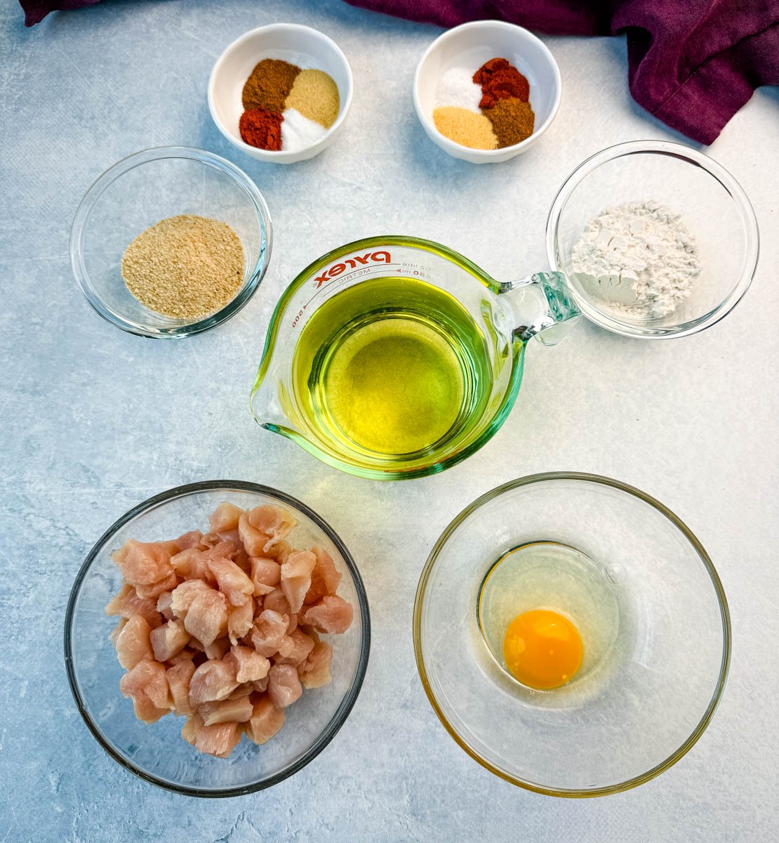 Overhead view of individual ingredients for fried chicken: chopped raw chicken, an egg, flour, breadcrumbs, seasonings, and oil.