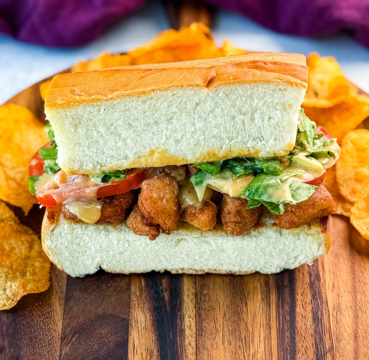 Front-facing shot of a stacked crispy chicken po' boy showing the golden chicken, fresh tomato slices, lettuce, and sauce inside a fluffy hoagie roll, with chips in the background.