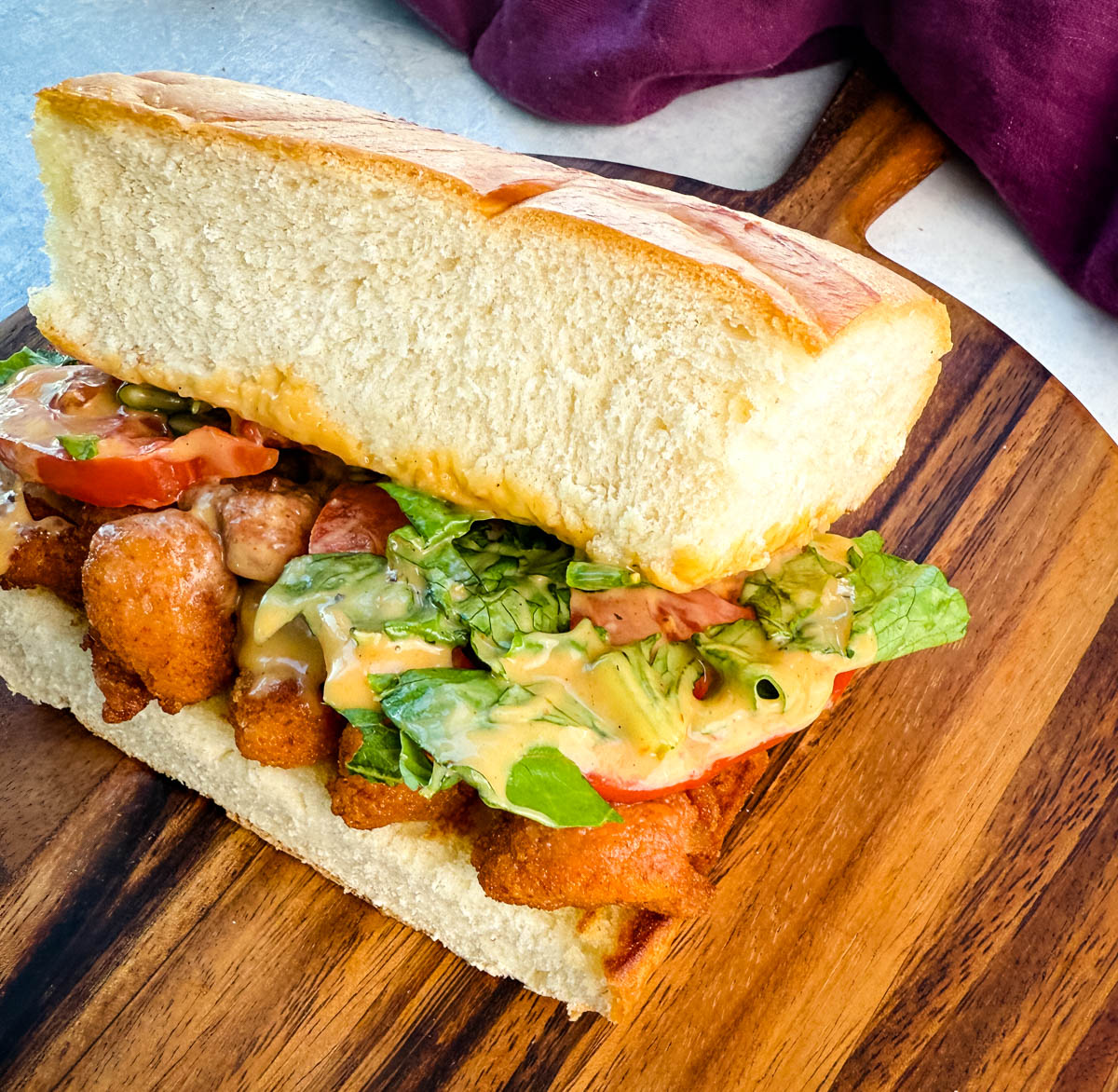 Angled view of a crispy chicken po' boy sandwich loaded with toppings and served with seasoned chips, resting on a wooden serving board.