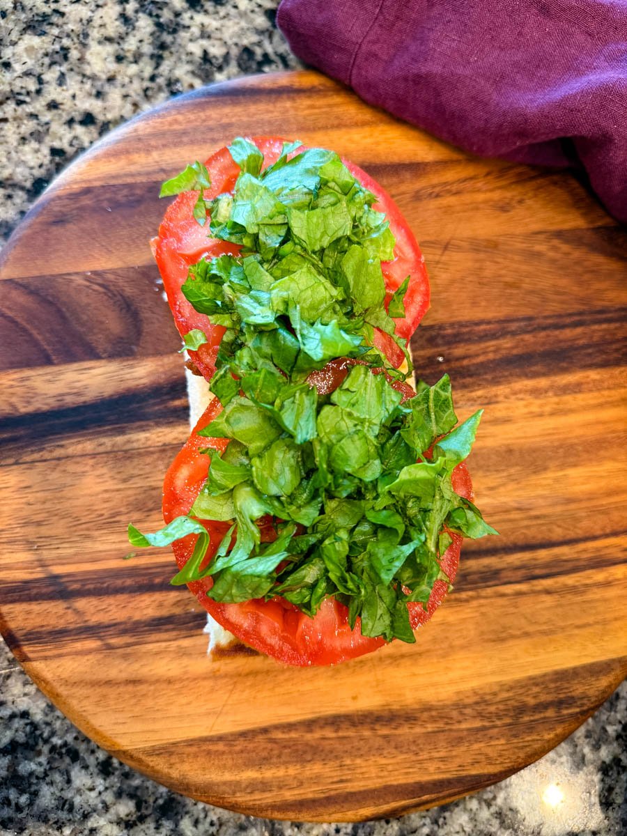 Overhead shot of a sandwich roll topped with fresh tomato slices and shredded romaine lettuce, on a wooden board with a purple napkin nearby.