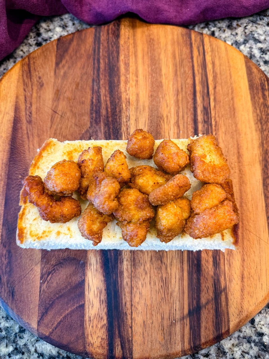 Top-down image showing the bottom half of a sandwich roll loaded with golden, crispy chicken pieces on a wooden board with a granite countertop background.