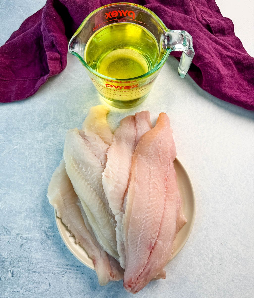 A small plate with raw catfish fillets stacked on top, next to a glass measuring cup filled with oil. A deep purple cloth napkin is in the background on a light blue surface.
