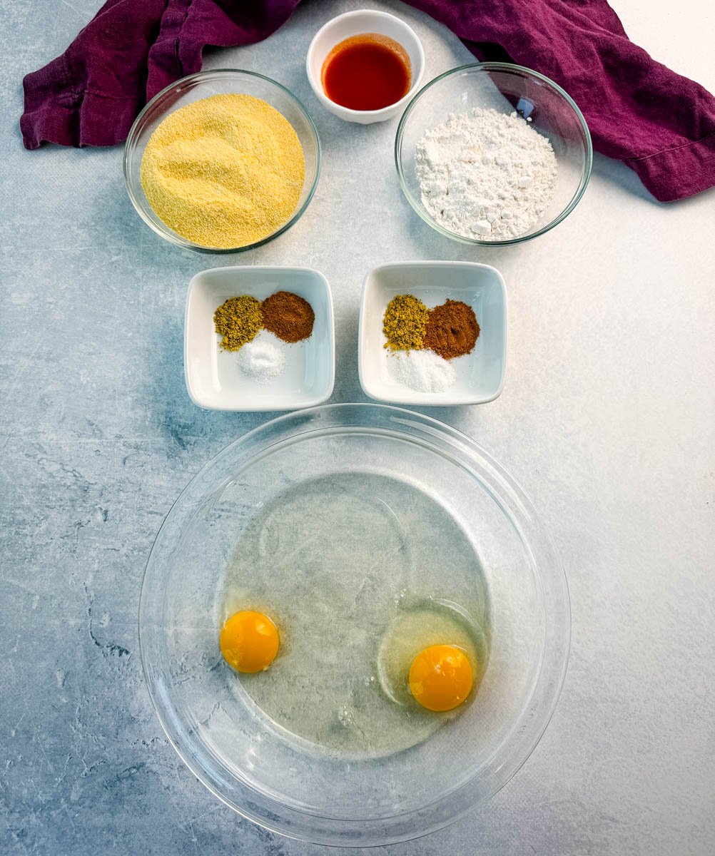 Flat lay of fish fry ingredients: cornmeal, flour, hot sauce, spices, and two cracked eggs in a clear bowl, all arranged on a light blue surface with a purple napkin.