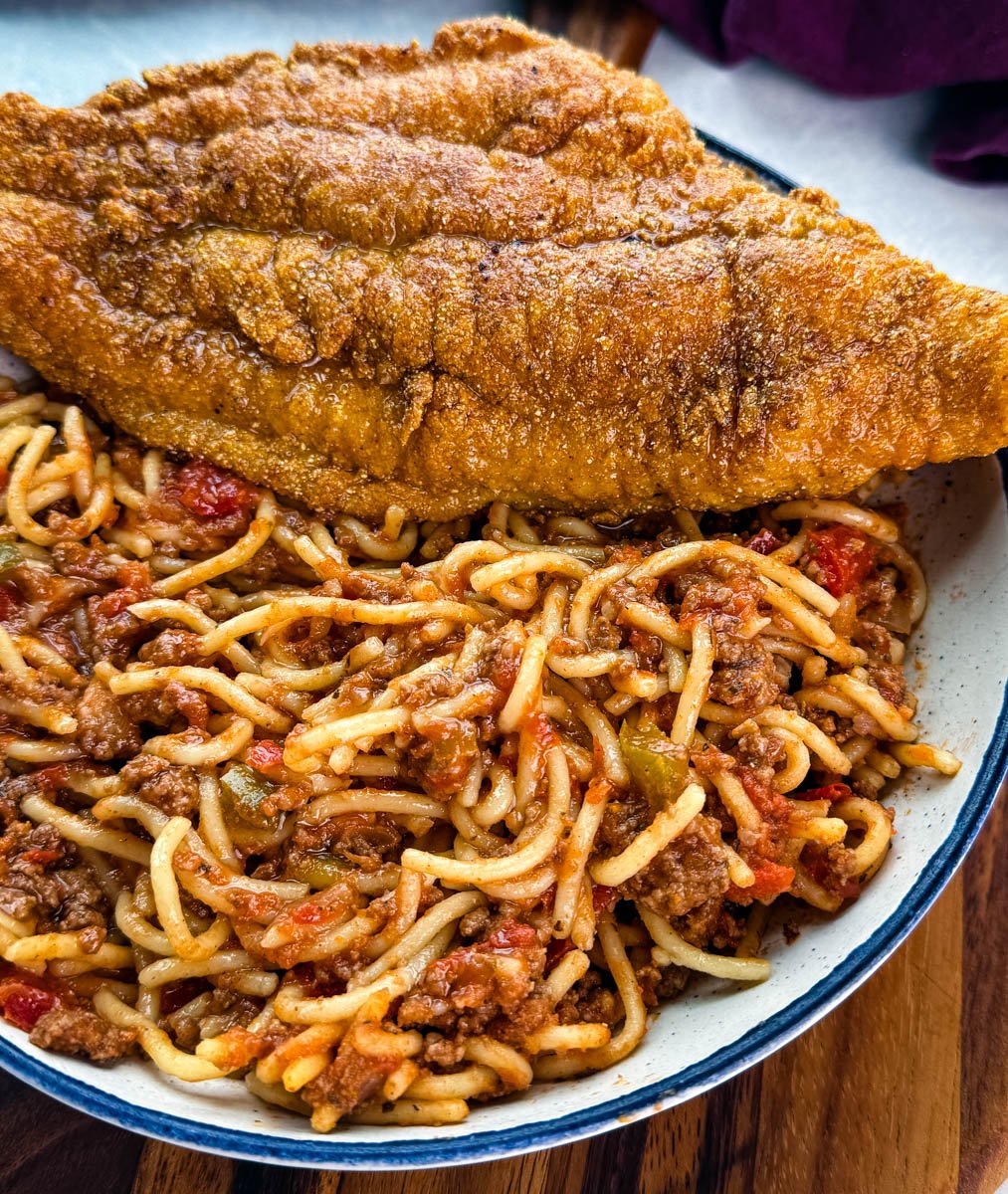 Close-up of a crispy fried catfish fillet resting on top of a bowl of spaghetti with a rich, chunky meat sauce.