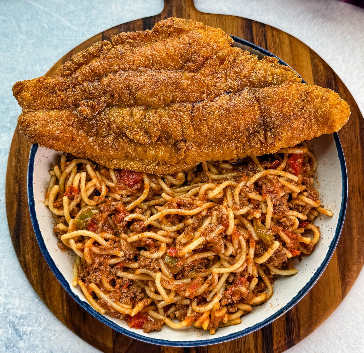 Overhead view of a serving of fried catfish placed over a bowl of spaghetti meat sauce, displayed on a wooden cutting board.