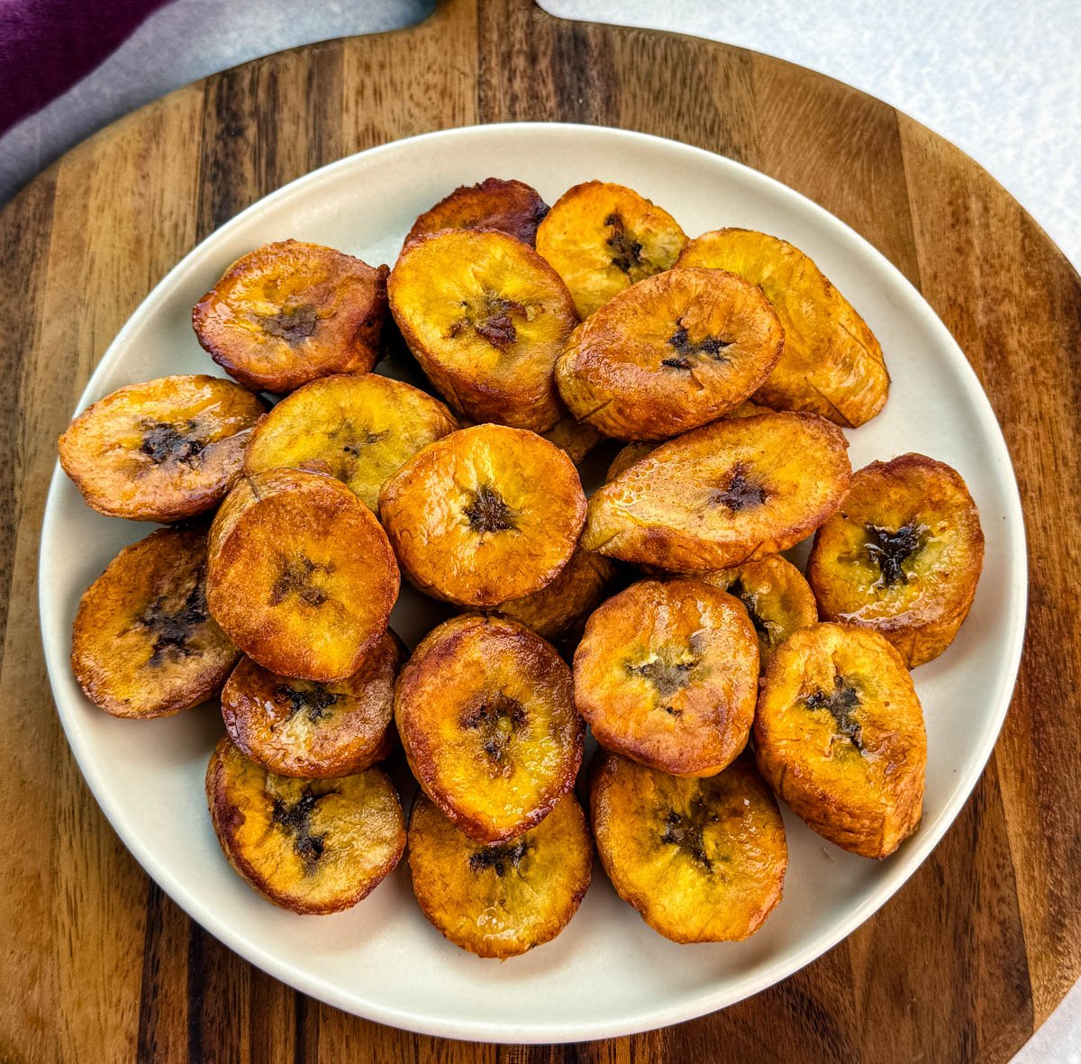 Plate of golden, crispy fried plantains stacked on a cream-colored plate set over a wooden board.