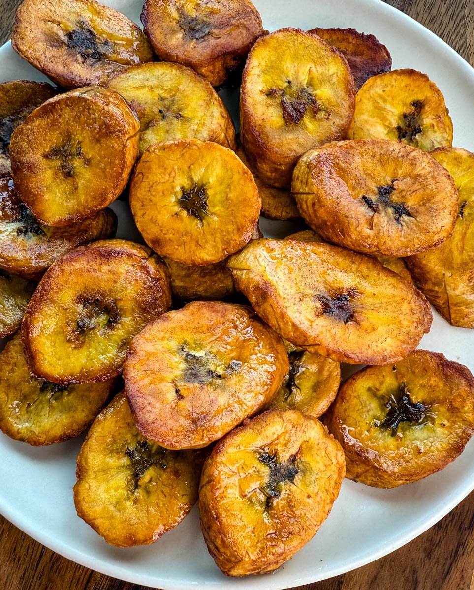 Close-up of perfectly golden-brown fried plantains stacked on a plate, glistening with oil.