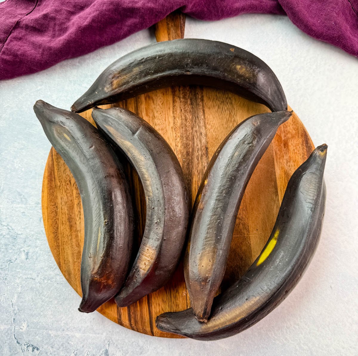 Fully ripened, dark black plantains on a round wooden board with a purple cloth.