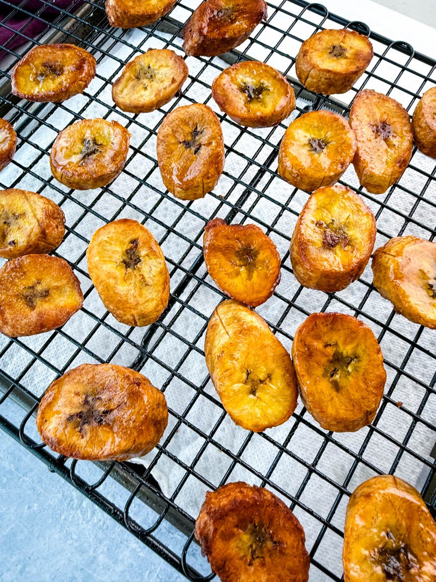 Overhead view of crispy fried plantains evenly spaced on a cooling rack with a slight sheen of oil.