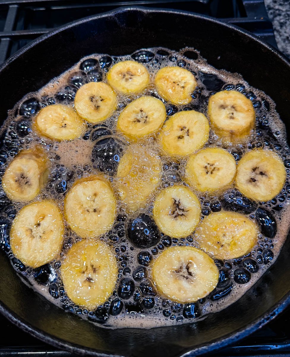 Raw plantain slices bubbling in oil in a black skillet, showing the start of the frying process.