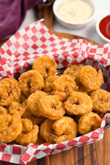 Front-focused shot of crispy fried shrimp piled high in a checkered basket, with sauce cups slightly out of focus in the background.